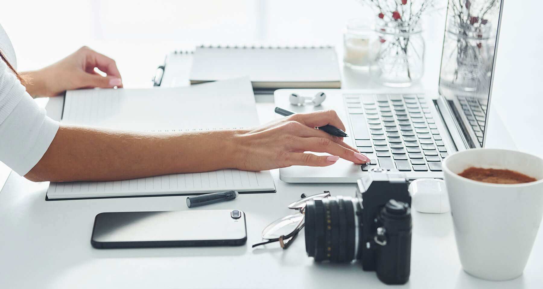 Woman working on content on laptop with camera notepad and iphone on desk